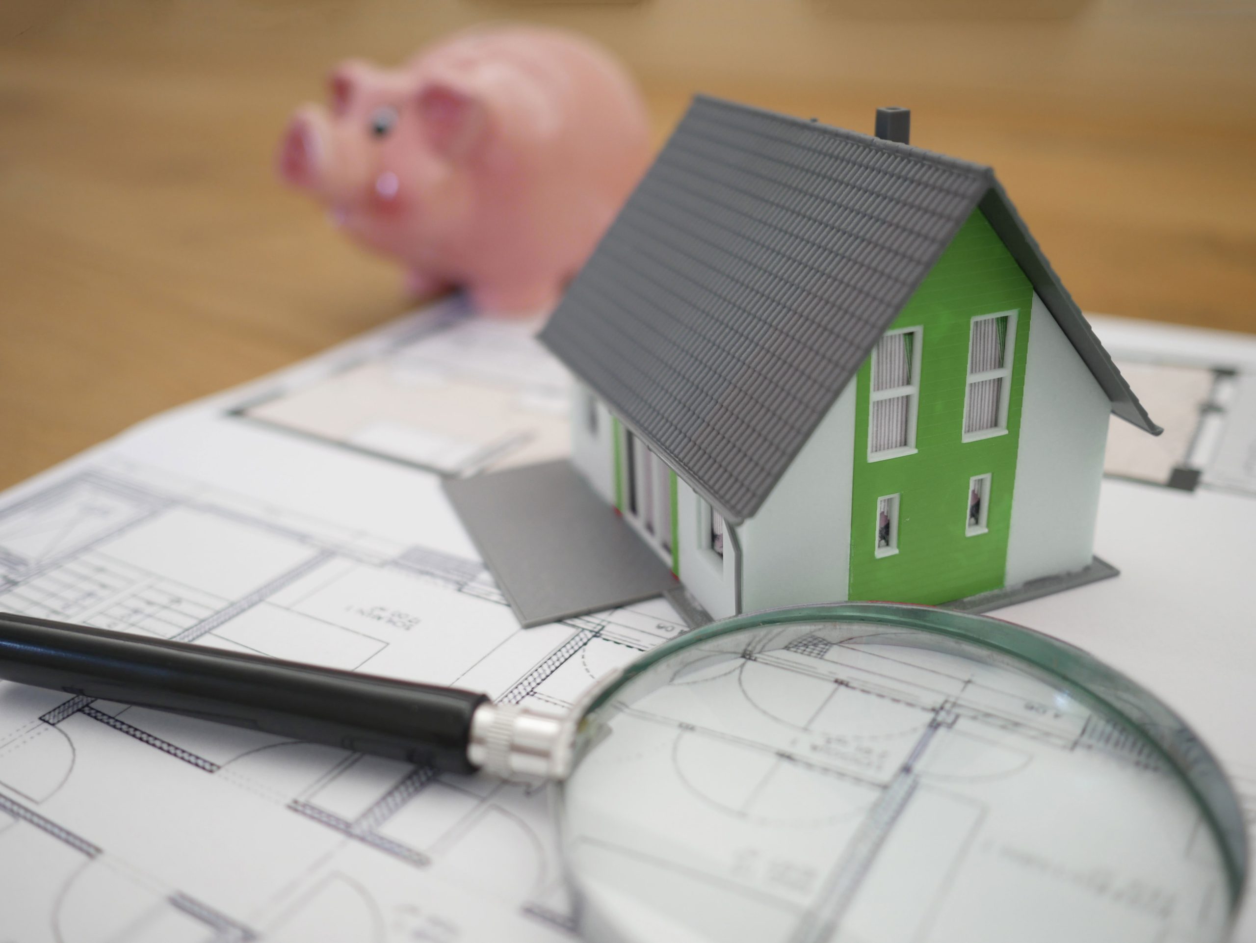 a small plastic green house with a magnifying glass and piggy bank beside it, sitting atop a blueprint plan of a house