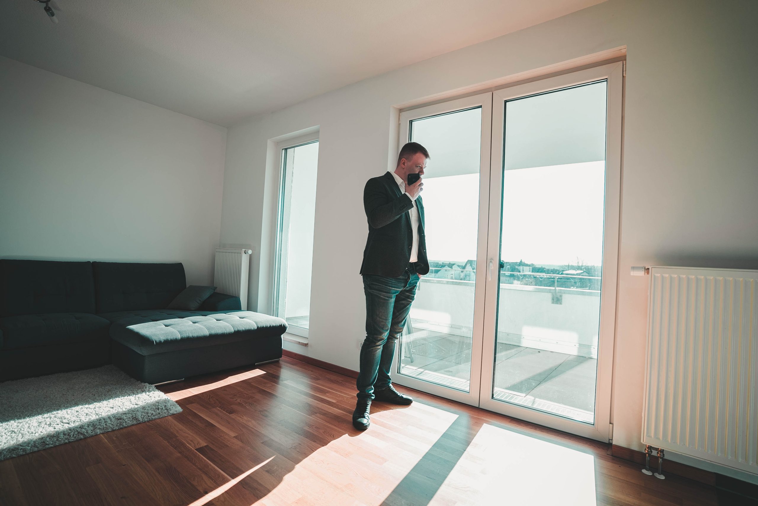 a business man on his mobile phone standing by glass double doors leading to a balcony inside a living room with a large black couch